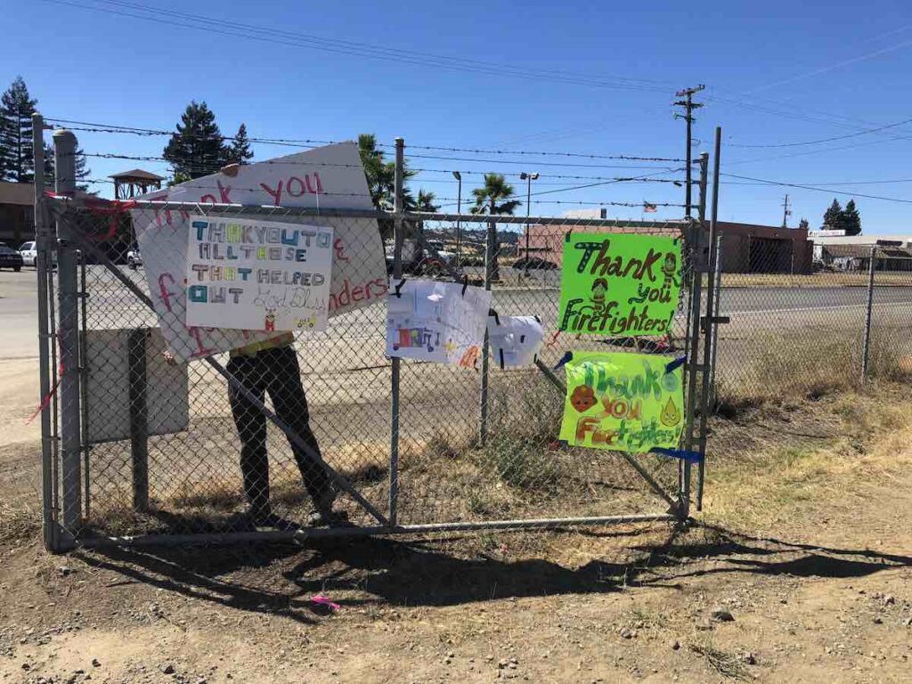 A person hangs up hand drawn signs saying "thank you firefighters" on a chain link fence.